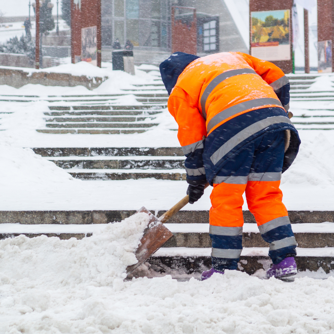 A worker in an orange and black jacket shovels snow on a snowy staircase during a winter storm.