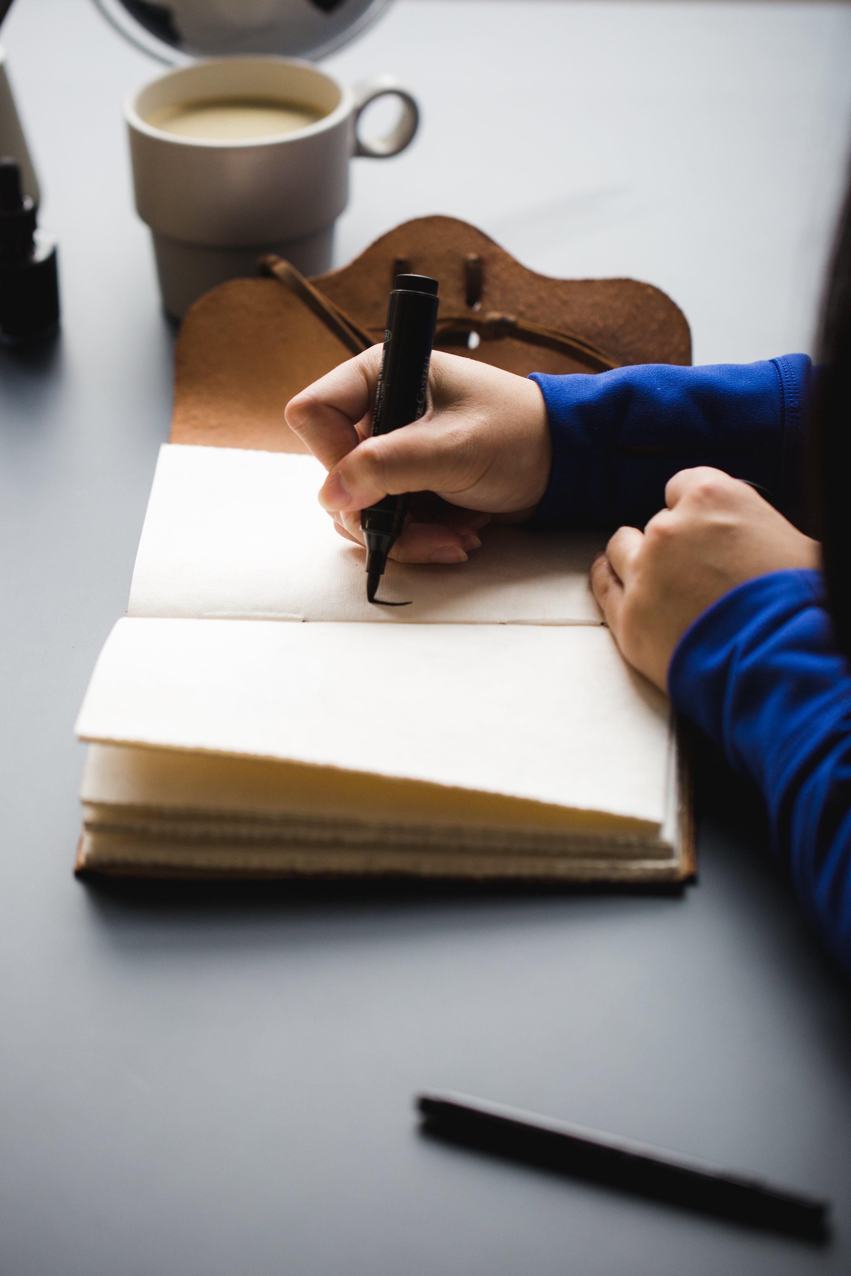 A person writing in a journal with a pen, surrounded by a cup and other stationery on a table.