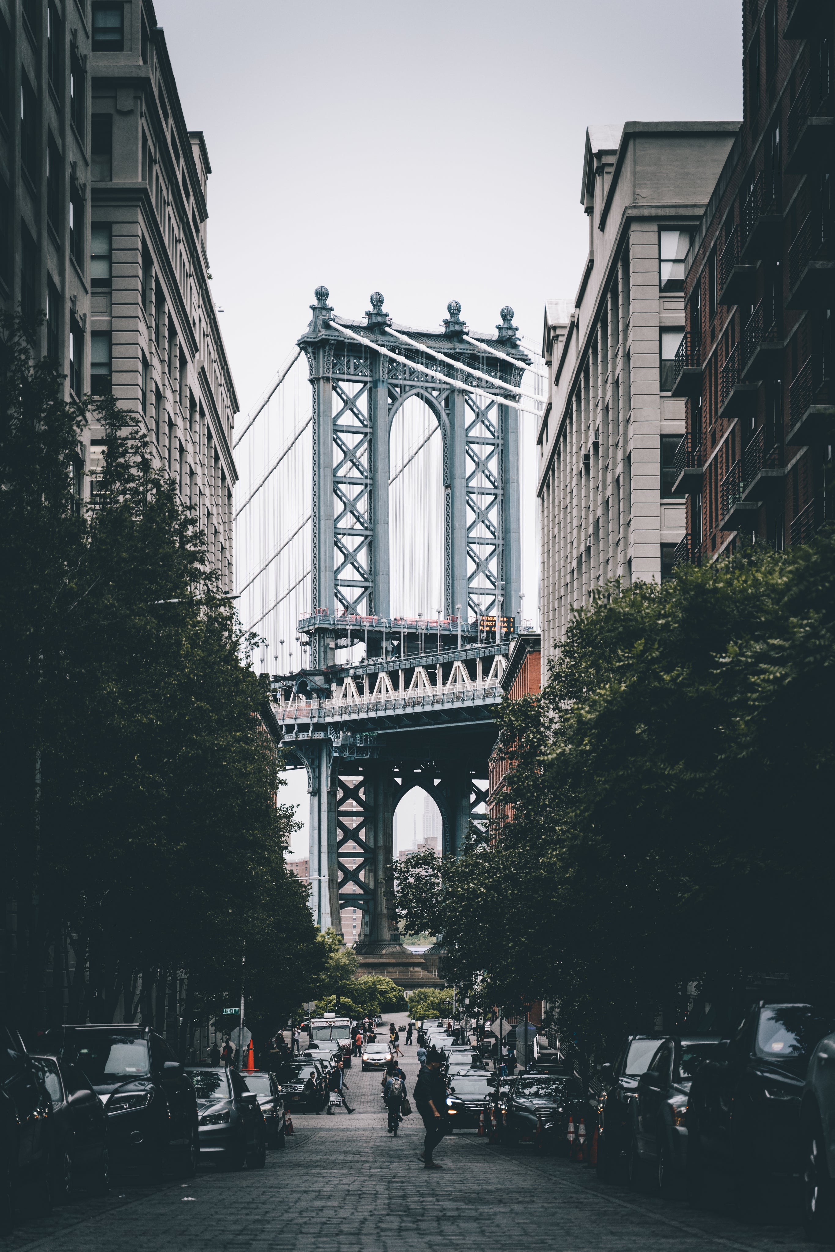 The image captures a view of the Manhattan Bridge as seen from a street lined with trees and parked cars.