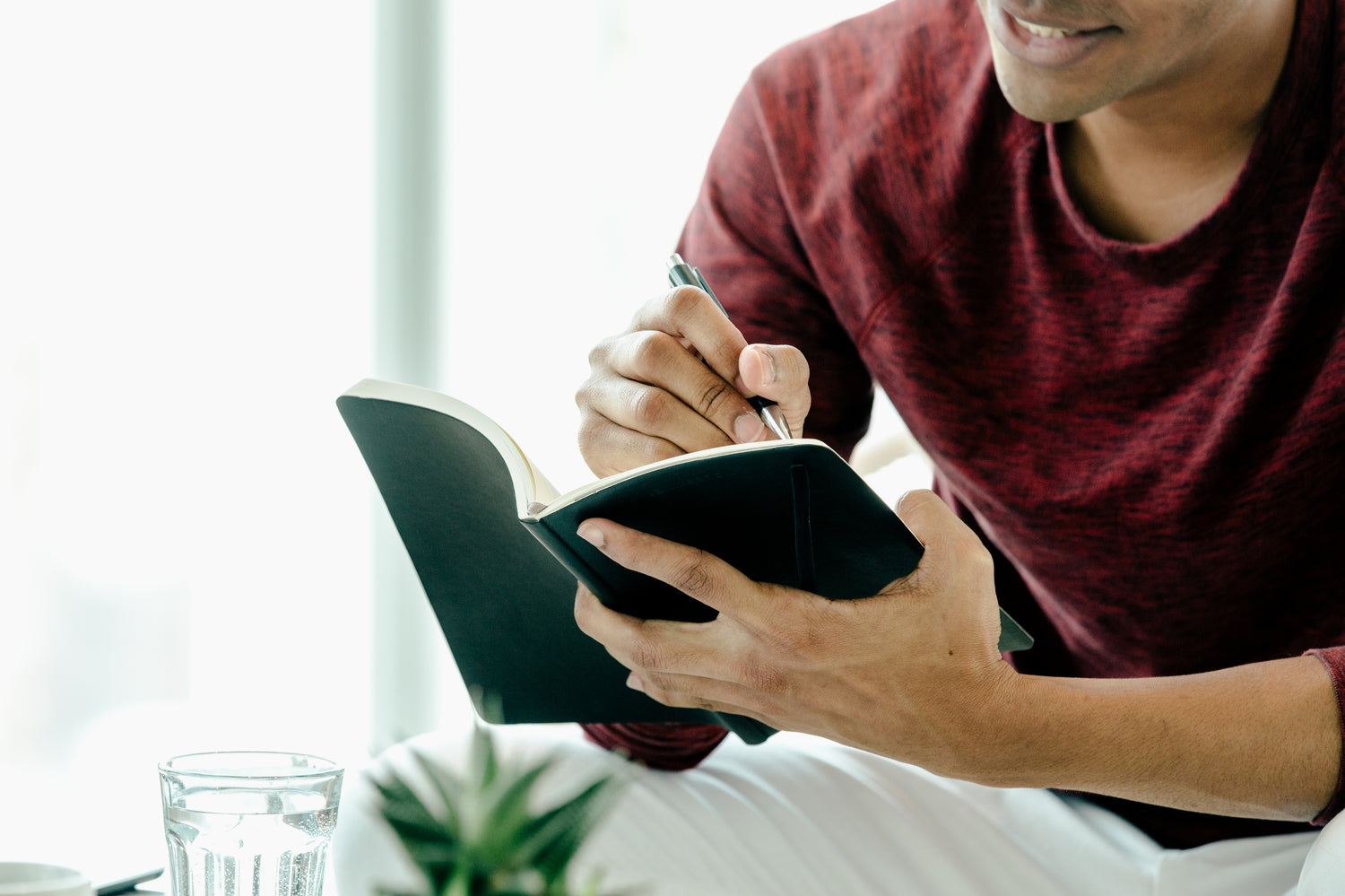 A person writing in a notebook while sitting at a table, with a glass of water nearby.