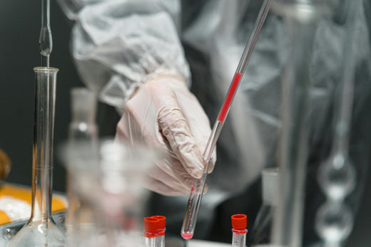 A gloved hand holds a test tube filled with red liquid, surrounded by various laboratory glassware on a table.
