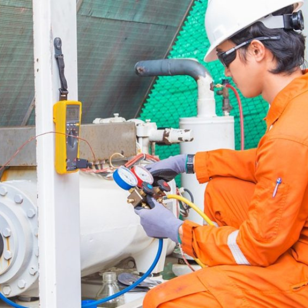 A technician in an orange jumpsuit and hard hat uses a multimeter on machinery, wearing safety goggles.