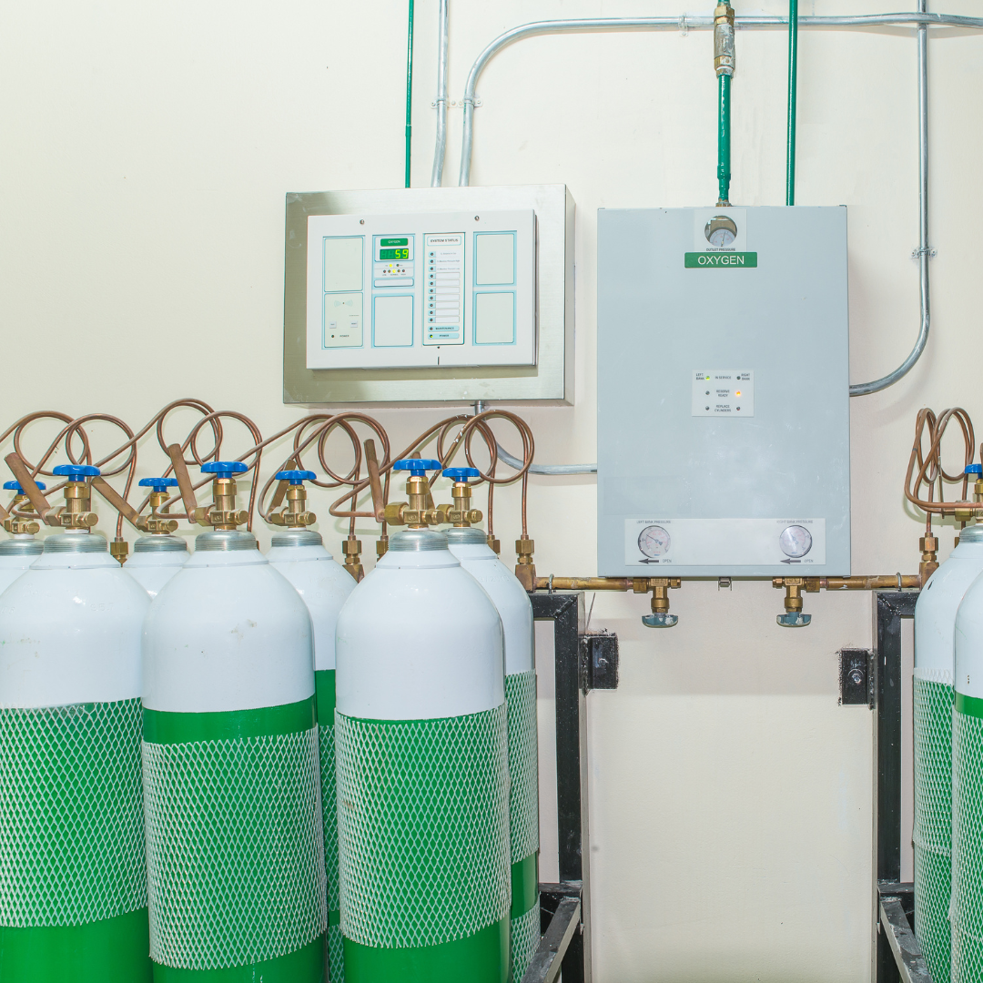 A row of medical oxygen tanks with green and white markings next to an oxygen control panel.
