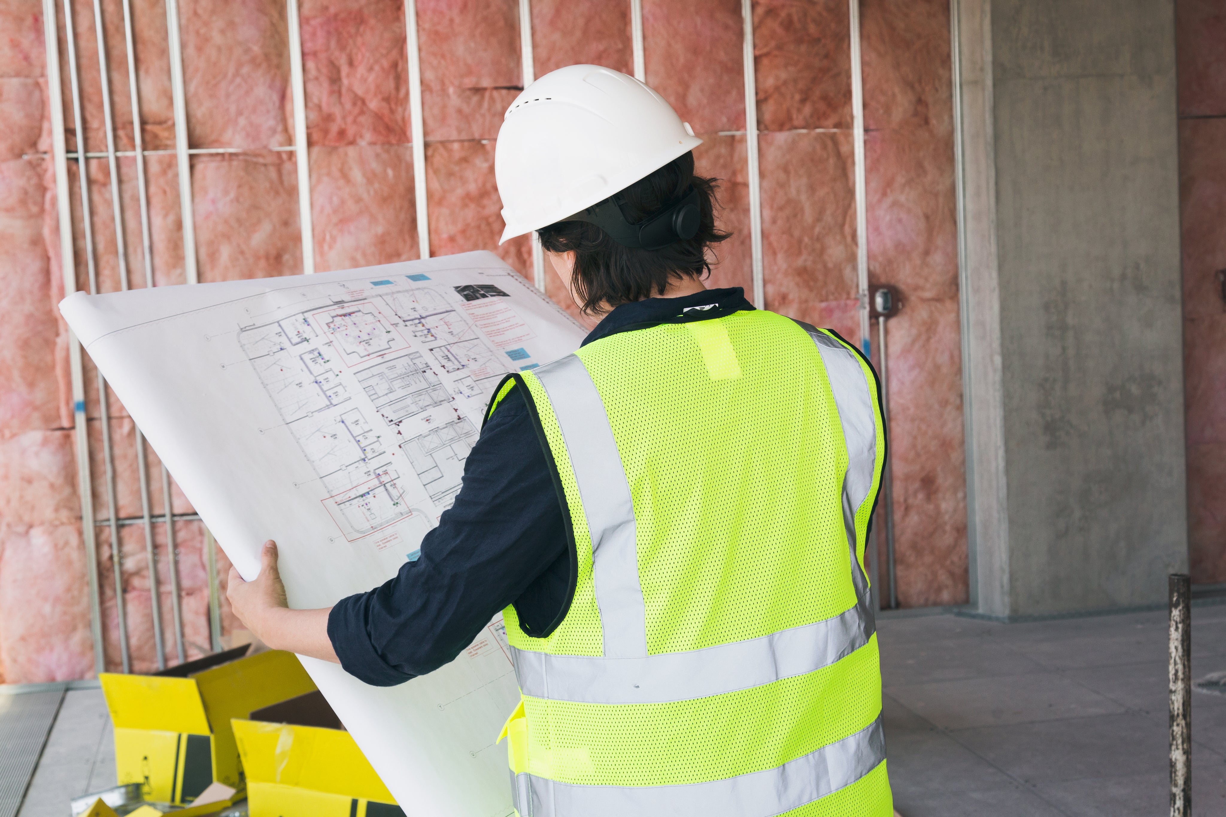 A construction worker in a hard hat and reflective vest examining blueprints at a construction site.