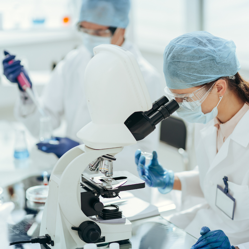 A laboratory scene featuring two scientists in white lab coats, wearing face masks and gloves.