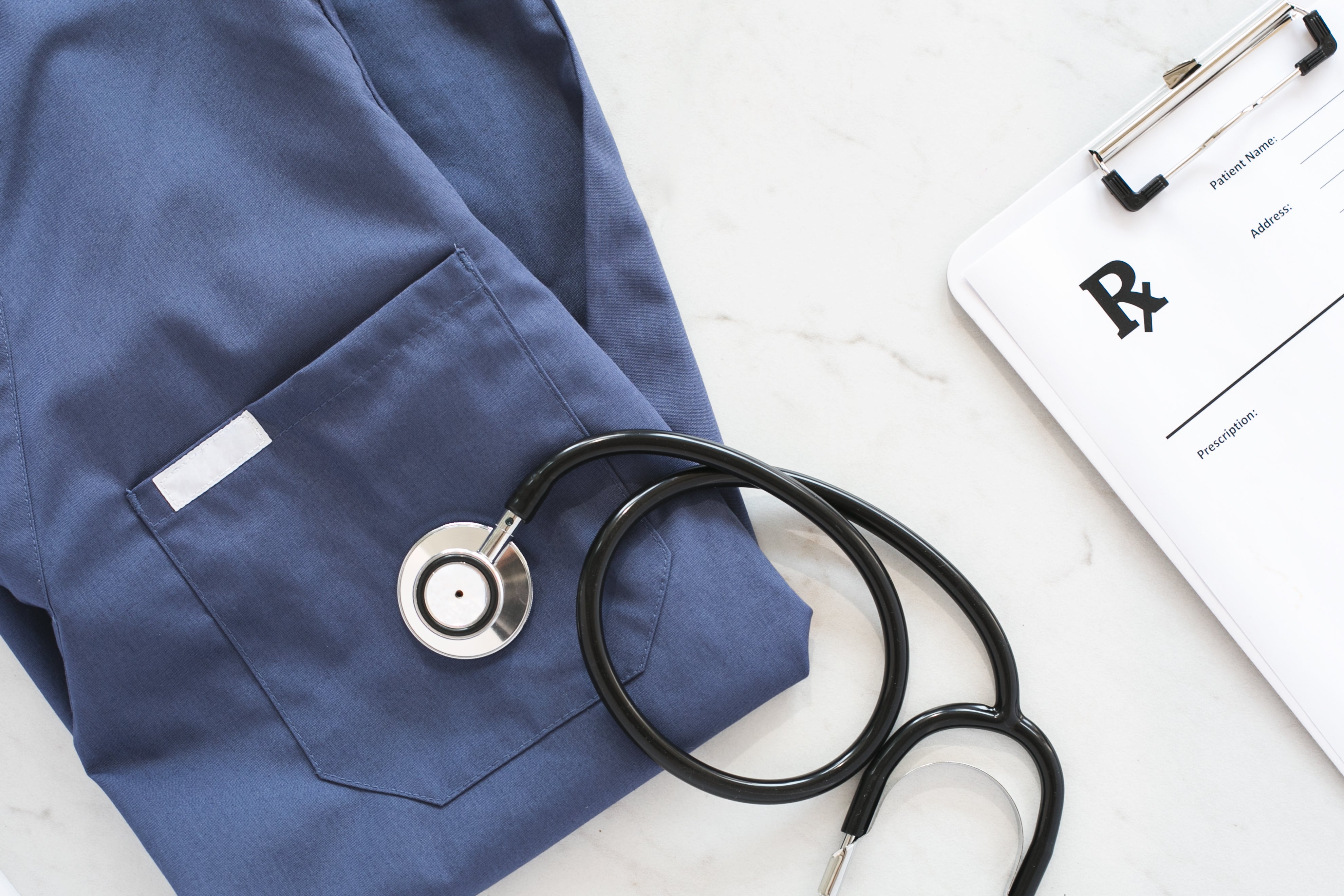 The image features a close-up of a navy blue medical top, a black stethoscope resting on it, and a clipboard with a prescription pad partially visible.